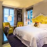Hotel room with a king bed featuring a bright yellow headboard, two green armchairs, and a desk.