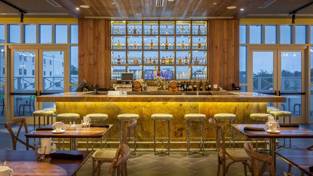 A brightly lit bar with a stone counter, backlit shelves of liquor bottles, and rows of stools, flanked by floor-to-ceiling windows.