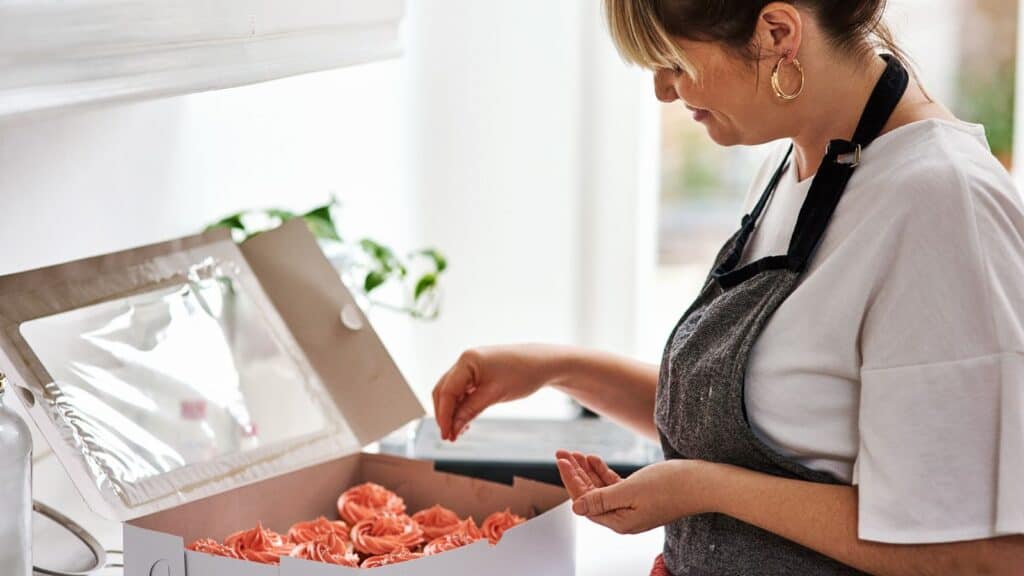 A woman in an apron, representing home cooks who legally sell food, places a pink-frosted cupcake into a bakery box.