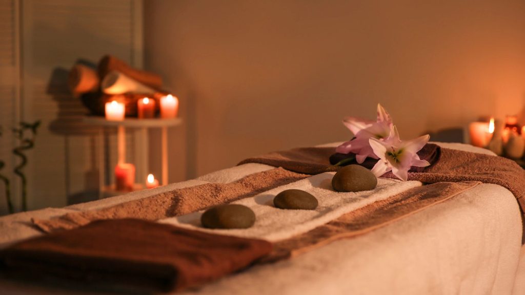 A massage table prepared with towels, three smooth stones, and a lily, with candles in the background.