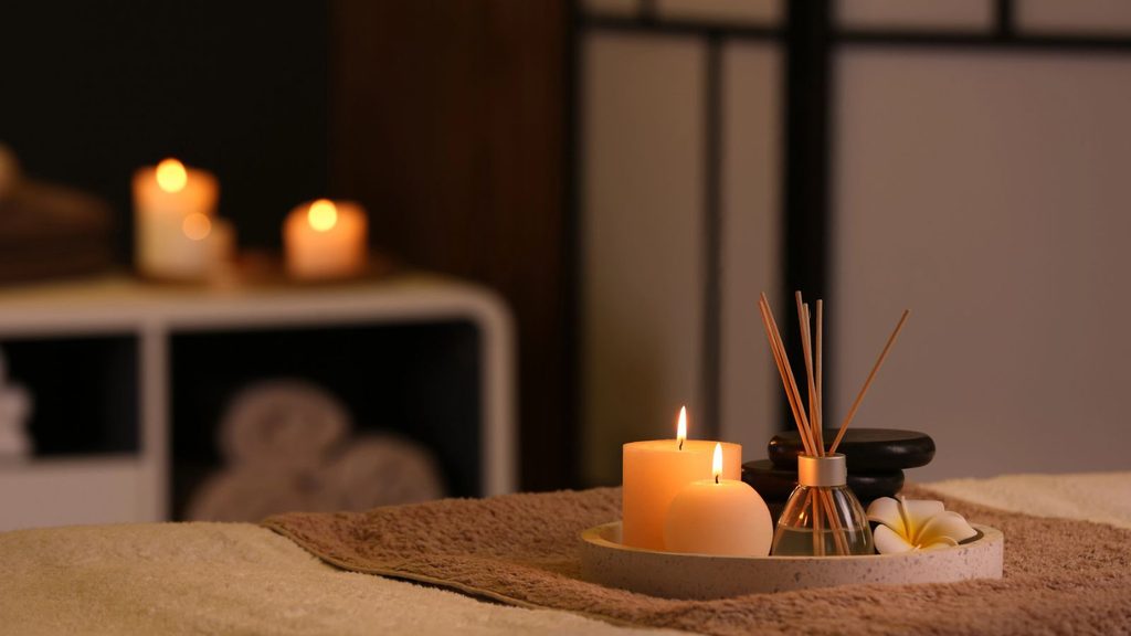 Two lit candles, a reed diffuser, black stones, and a flower on a tray on a massage table.