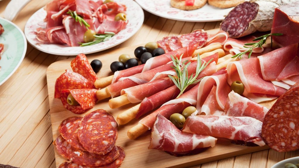 A wooden board topped with various cured meats, breadsticks, and olives, with additional plates of meat in the background.