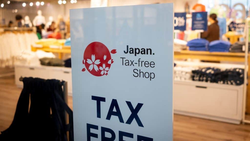 A white sign in a store reading &ldquo;Japan. Tax-free Shop&rdquo; with a red circle logo containing white cherry blossom designs.