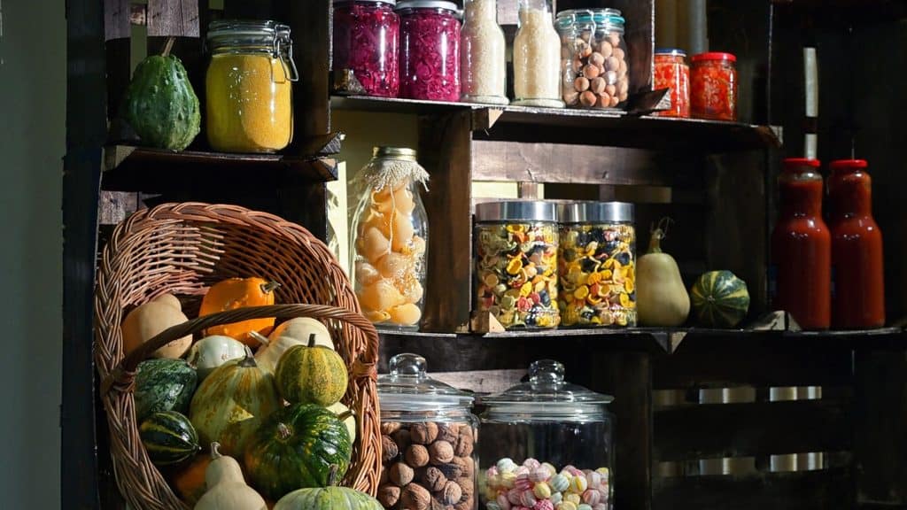 A rustic wooden shelving unit displaying glass jars of grains, pasta, nuts, and candies, alongside a wicker basket filled with various gourds and squash.