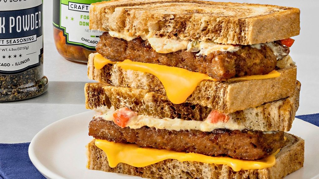 A double-decker sandwich with sausage patties, melted cheddar cheese, and red pepper spread on toasted bread, with seasoning jars in the background.