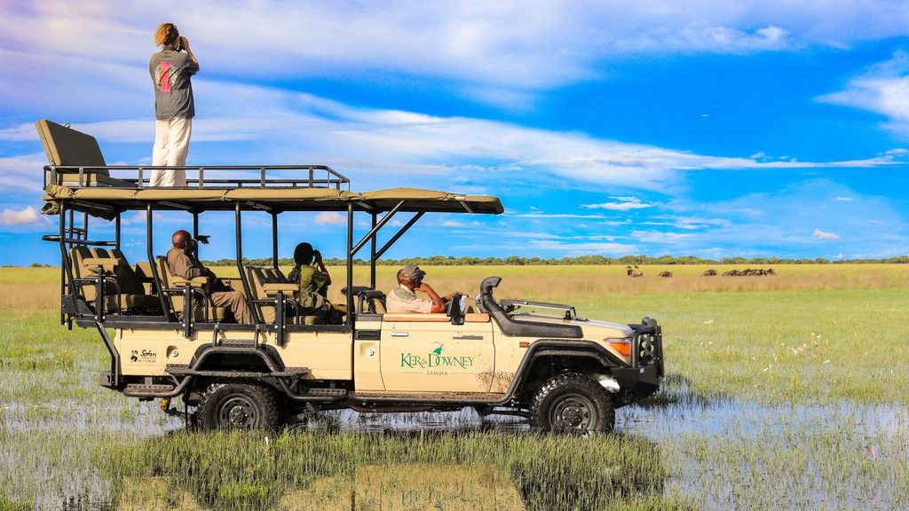 A safari vehicle driving through the shallow, grassy wetlands of Liuwa Plain National Park, with one person standing on the roof and others inside.