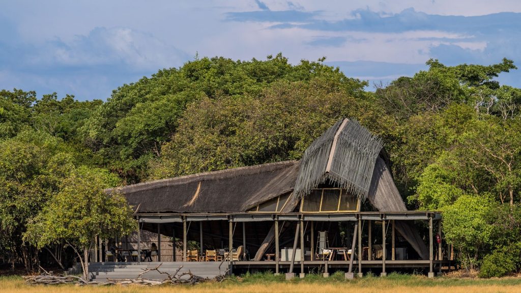 A large open-air lodge with a thatched roof, set among trees and surrounded by the tall grass of Liuwa Plain National Park.
