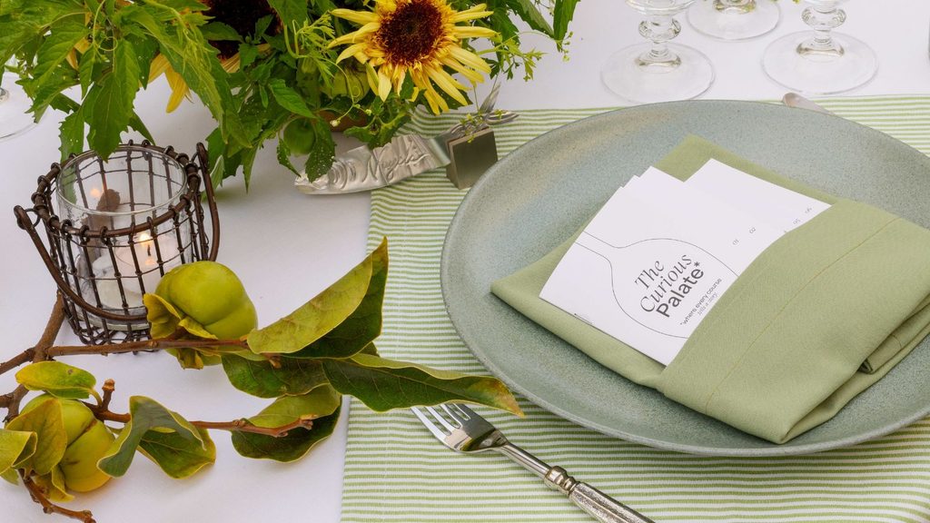 A Kimpton holiday cocktail place setting with a green plate, a green napkin, and a menu card labeled “The Curious Palate,” next to a wire candle holder and a sprig of persimmons.