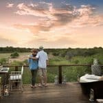 A couple stands on an elevated wooden deck overlooking a savanna at sunset.