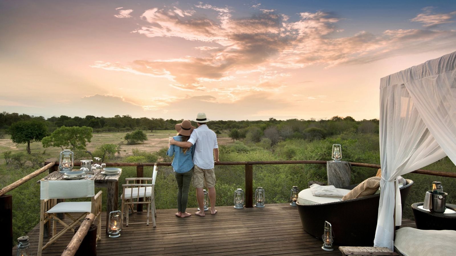 A couple stands on an elevated wooden deck overlooking a savanna at sunset.