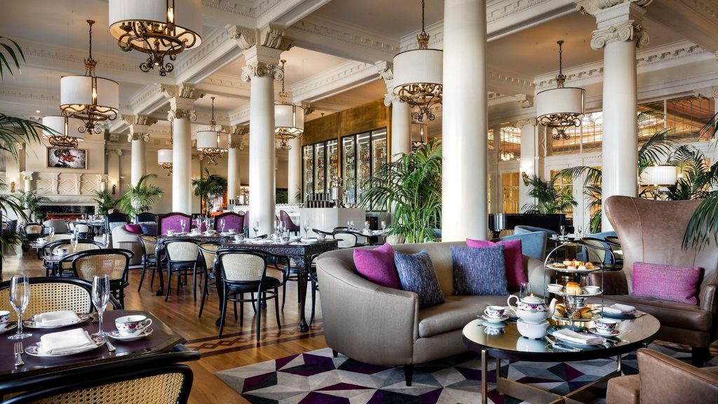 A spacious Fairmont Empress lounge with white columns, chandeliers, dining tables with patterned chairs, and a curved sofa set with tea service.