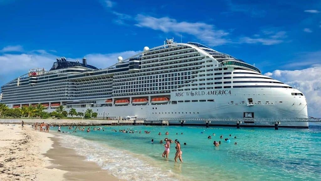 The MSC World America cruise ship docked at a pier beside a sandy beach with people swimming in the turquoise water.