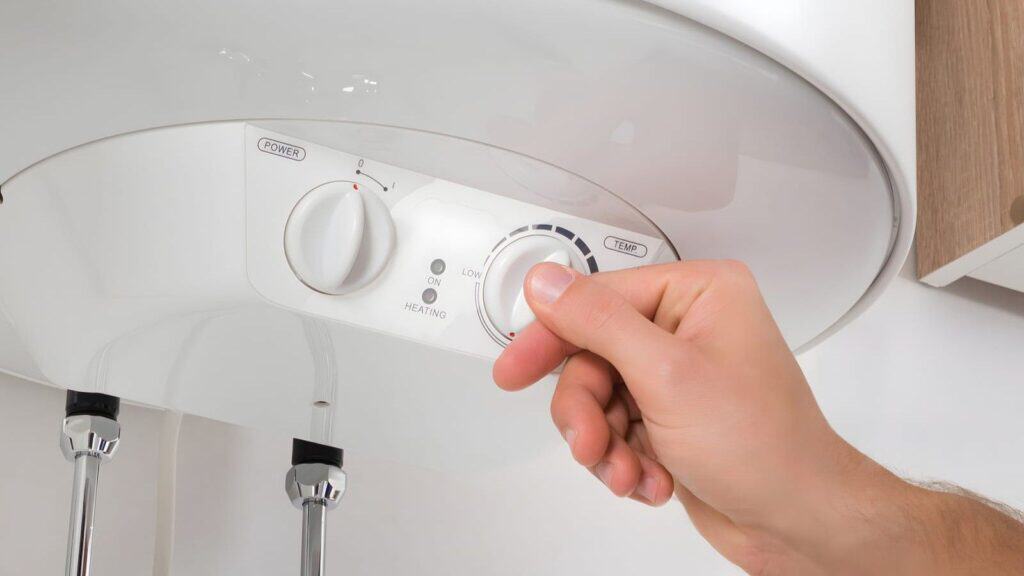 A hand adjusting the temperature dial on a white electric water heater to help weatherproof the home before a cold snap.