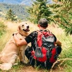A hiker with a red backpack sits on a grassy hill with their arm around a golden retriever looking back at them.