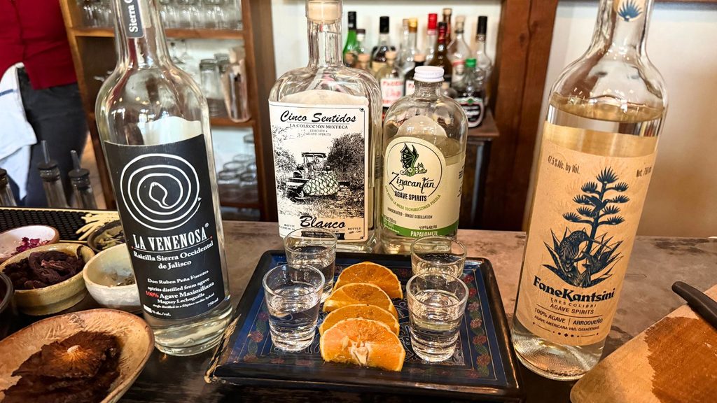 Four bottles of mezcal and raicilla arranged behind a tray of shot glasses and orange slices at a Tucson city bar.