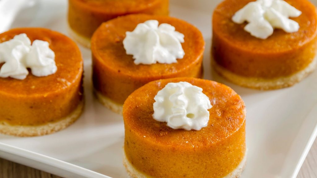 Five small, round pumpkin tarts topped with dollops of whipped cream on a white plate, perfect for halloween shopping.