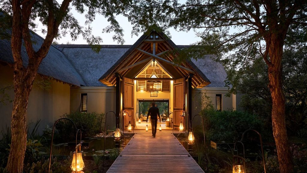 A person walks down a wooden footbridge towards the entrance of a lodge building, lit by lanterns.