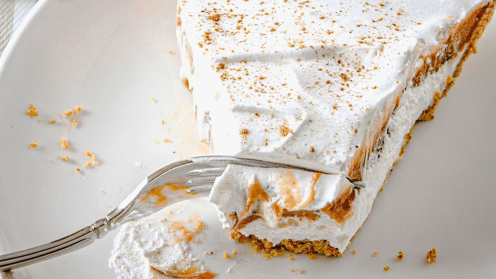 A fork taking a bite of a slice of National Pumpkin Cheesecake Day pie topped with whipped cream and spices on a white plate.