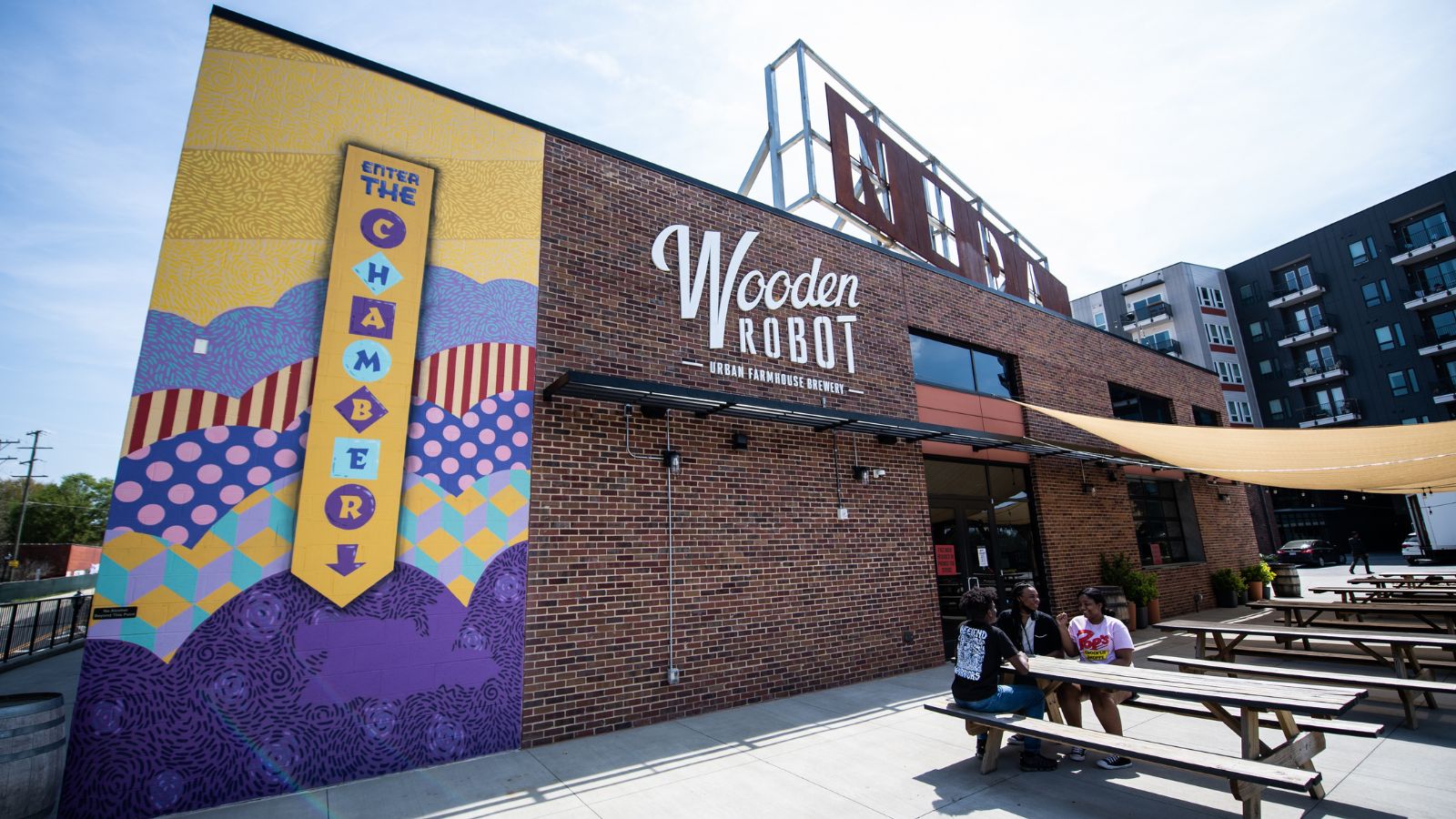 The exterior of Wooden Robot Urban Farmhouse Brewery, one of the top Charlotte breweries, featuring a colorful mural and outdoor picnic tables with people sitting at one.