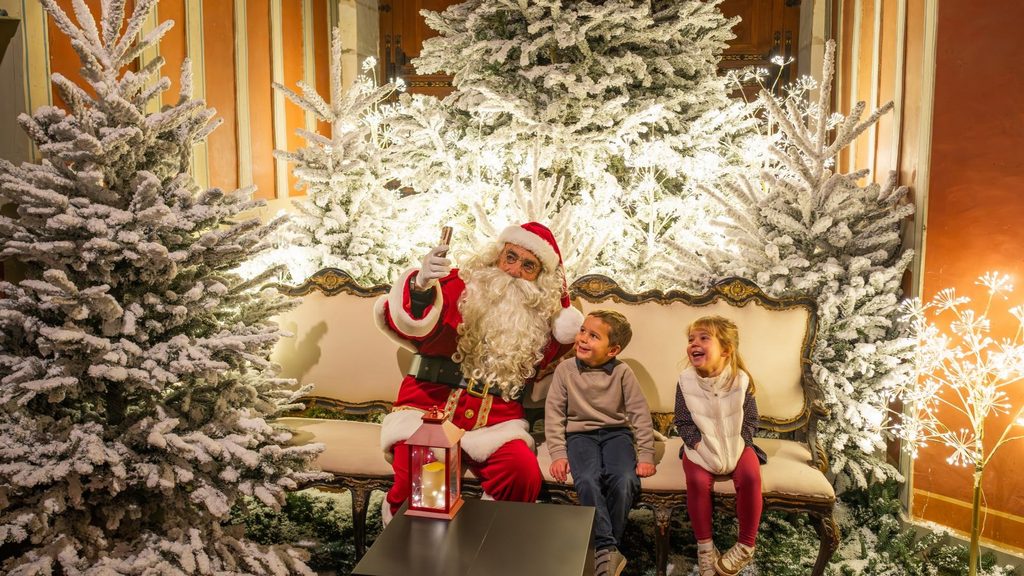Santa Claus takes a selfie with two children on a decorated bench surrounded by snowy Christmas trees, evoking Christmas in the land of ch&acirc;teaux.