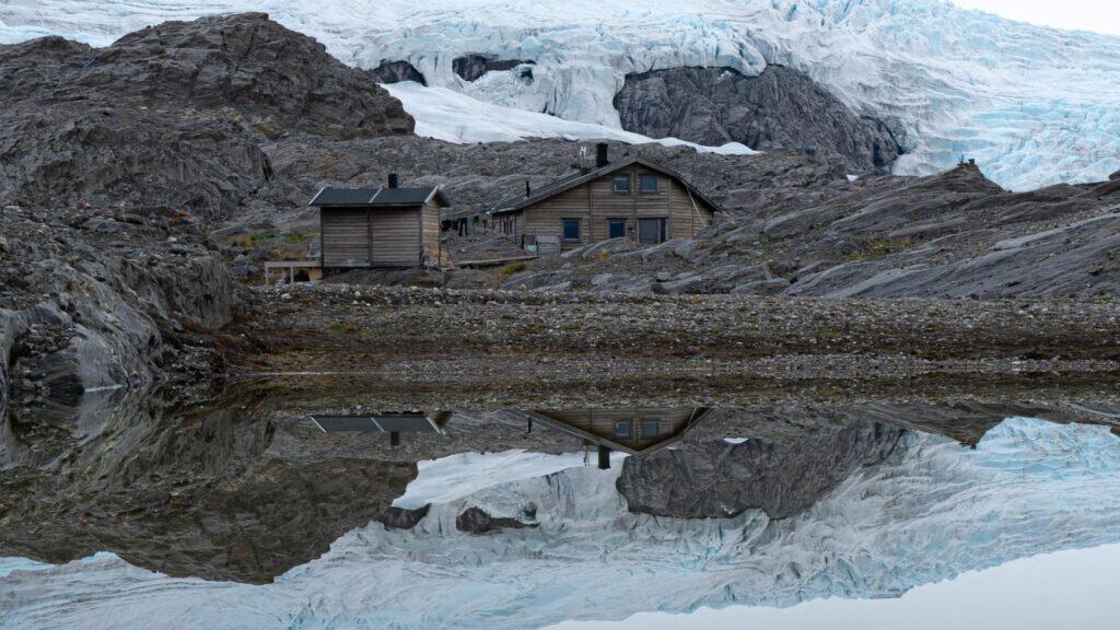 Two wooden cabins situated on a rocky slope in front of a glacier, reflected in the calm water of a lake during a Svalbard trip.