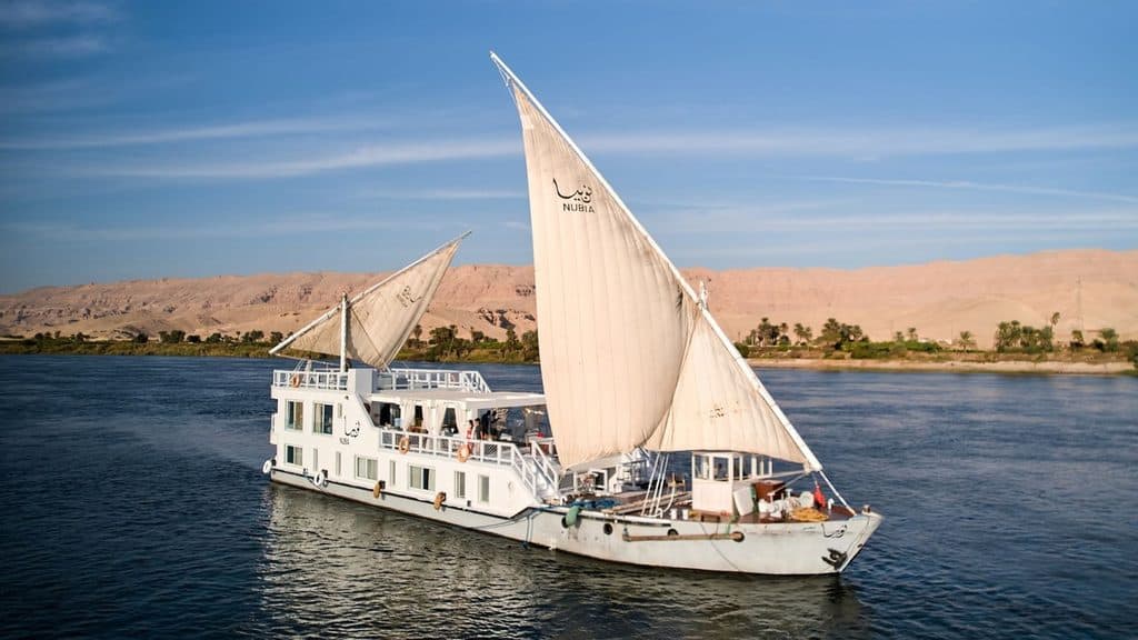 A white dahabiya sailing boat with two large sails on the Nile River, with arid cliffs in the background.