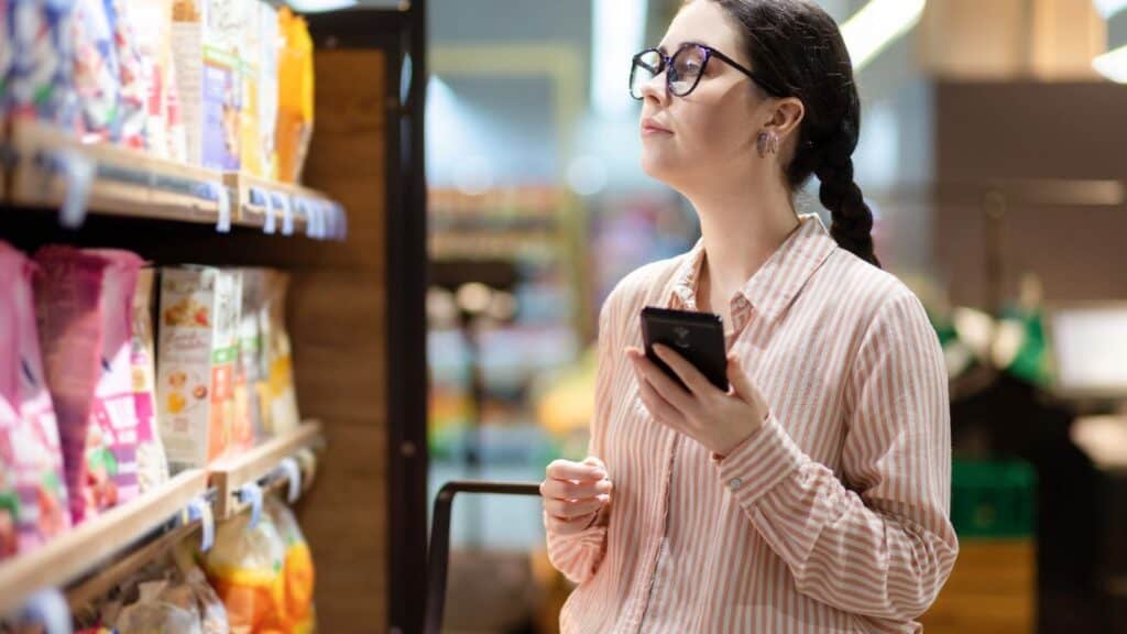 A woman wearing glasses and a striped shirt holds a smartphone while looking at products on a grocery store shelf.
