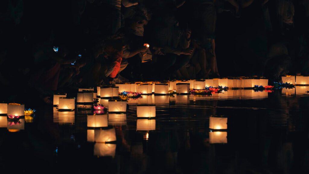 People crouching at the edge of a dark body of water, placing lit square paper lanterns onto the surface in a manner reminiscent of countries with Halloween-like traditions.