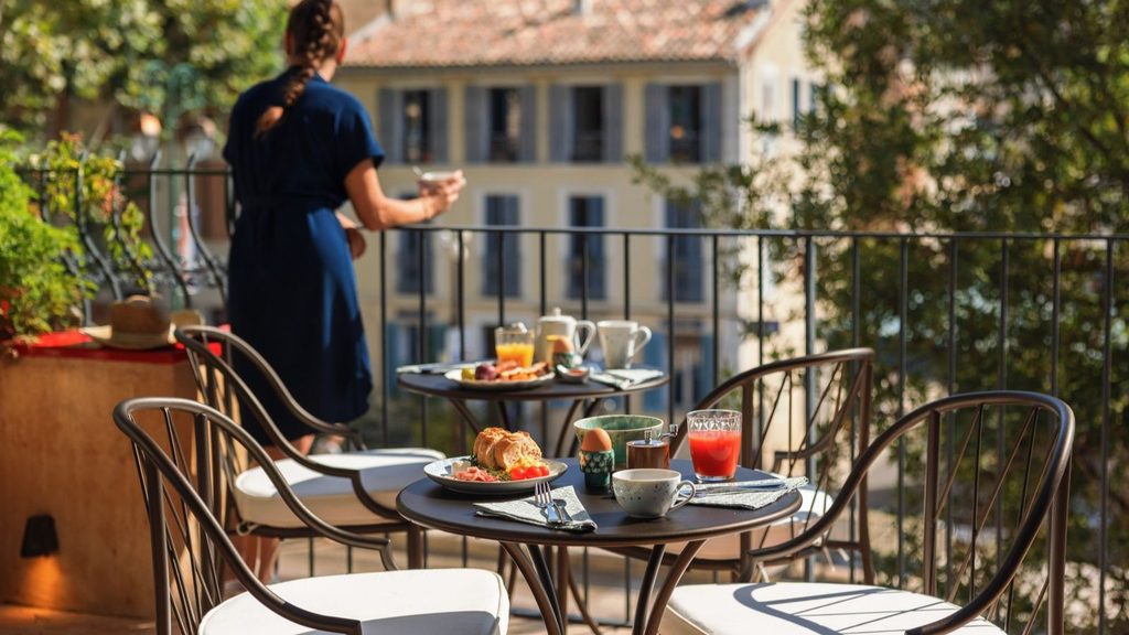 A balcony table set with breakfast items including bread, a soft-boiled egg, and juice, with a person standing by a second table in the background.