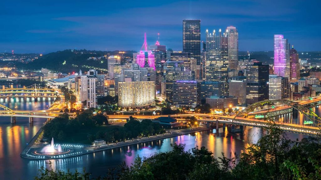 Pittsburgh city skyline at night with illuminated buildings and bridges over the river.