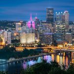 Pittsburgh city skyline at night with illuminated buildings and bridges over the river.