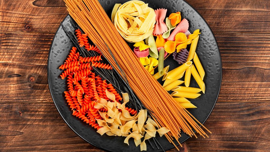 A black plate filled with various types of dry pasta, including spaghetti, fusilli, penne, and fettuccine, arranged on a dark wooden surface for National Pasta Day.