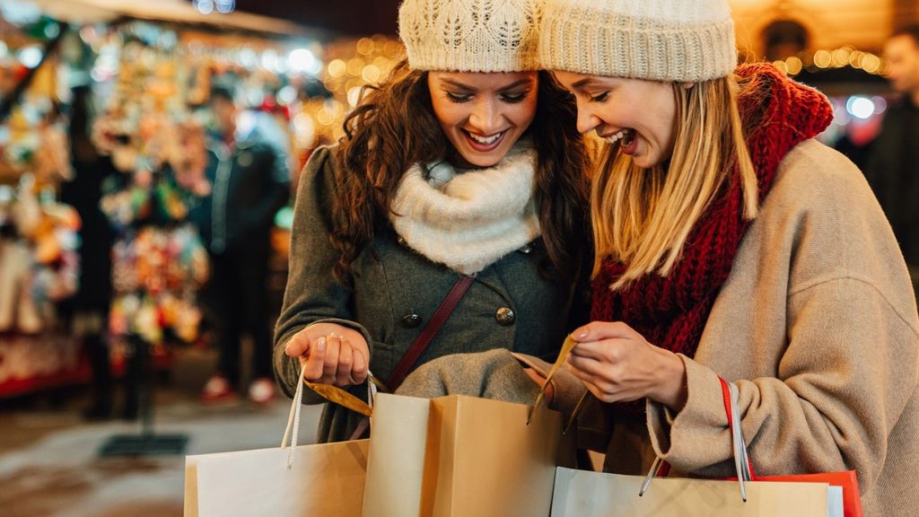 Two women in winter hats and scarves smiling as they look into a shopping bag while holiday shopping at a market.