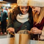 Two women in winter hats and scarves smiling as they look into a shopping bag while holiday shopping at a market.