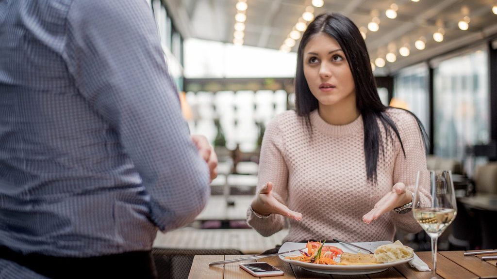 A woman gestures with her hands while talking to a person standing across from her at a restaurant table.