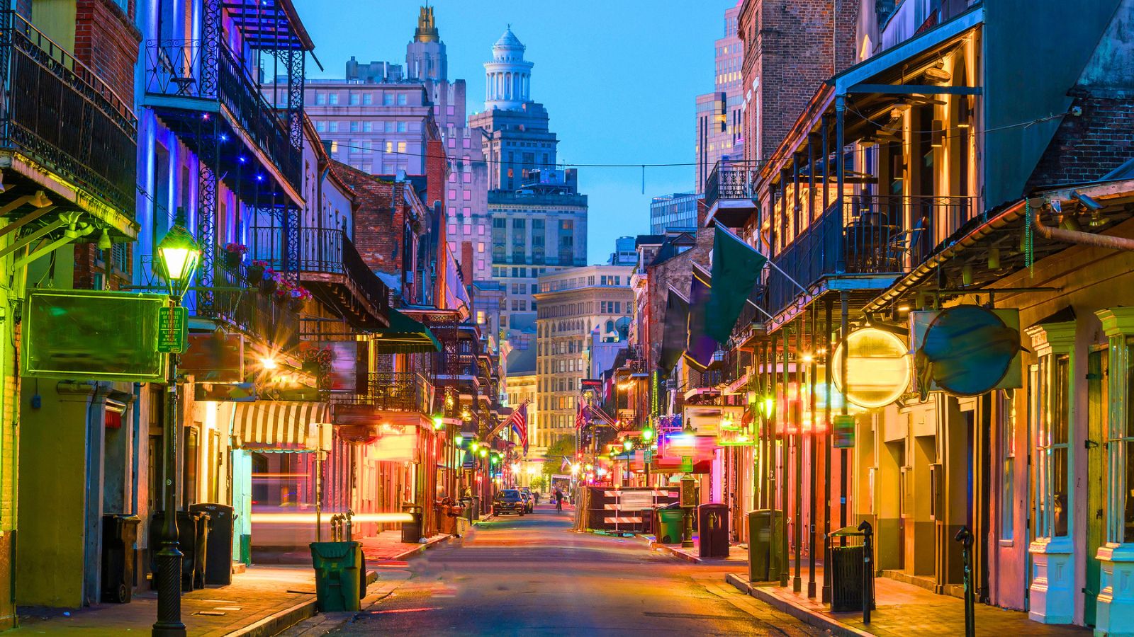 A narrow street in the French Quarter of New Orleans at twilight, perfect for fall travel, lined with multi-story buildings, wrought-iron balconies, and glowing storefronts.
