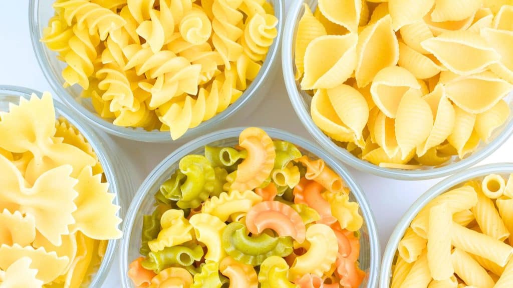 Four glass bowls filled with various shapes of uncooked pasta, including rotini, shells, farfalle, and multi-colored macaroni.