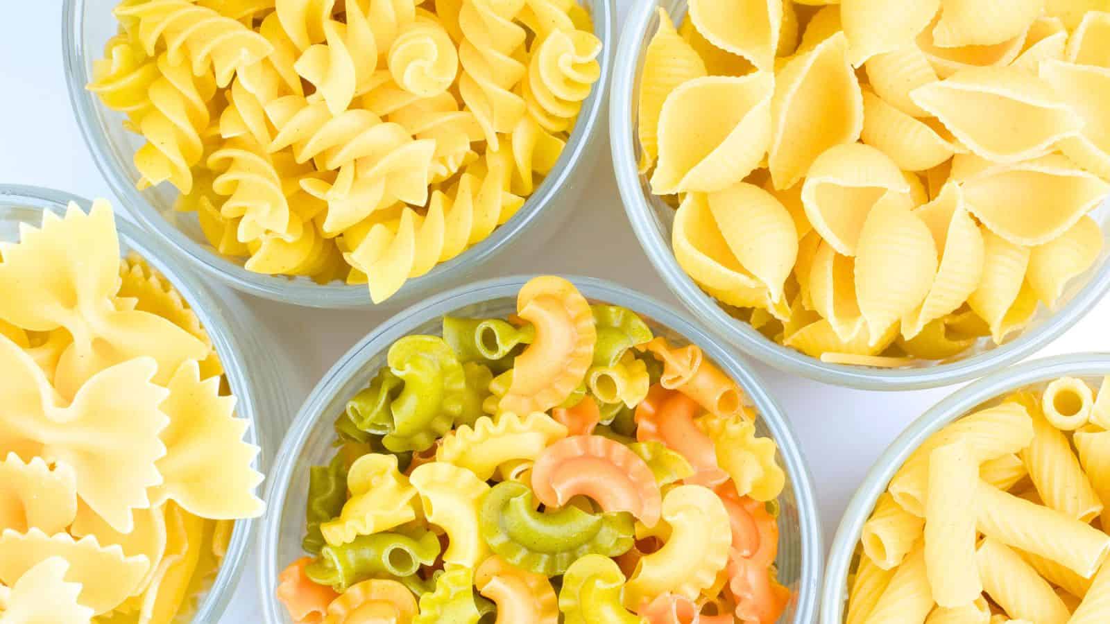 Four glass bowls filled with various shapes of uncooked pasta, including rotini, shells, farfalle, and multi-colored macaroni.