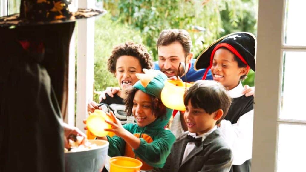 A group of children in Halloween costumes and an adult stand at a doorway with porch fixes before trick-or-treaters arrive, holding buckets to receive candy from someone in a witch hat.