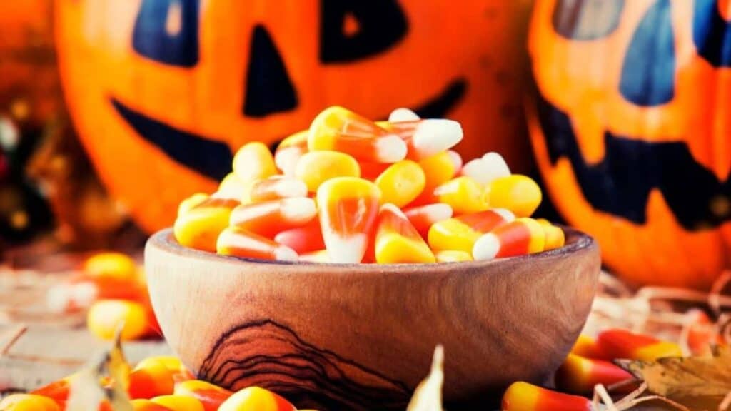 A wooden bowl filled with candy corn, one of the oldest Halloween candies, sits before two carved jack-o&rsquo;-lanterns.