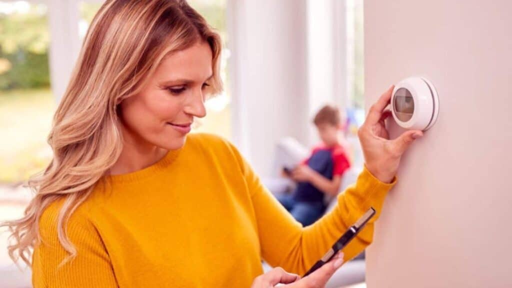 A woman in a yellow sweater adjusts a smart thermostat while holding a smartphone, debunking thermostat myths that won&rsquo;t save money.