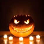 A jack-o’-lantern with a jagged, menacing face, illuminated from within and surrounded by five small, potential fire hazard candles on a wooden surface.