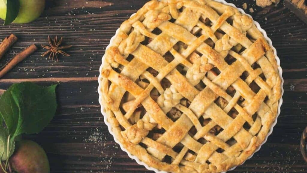 Lattice-topped apple pie in a white dish, surrounded by apples, cinnamon sticks, and star anise on a wooden surface for fall dessert recipes without pumpkin.
