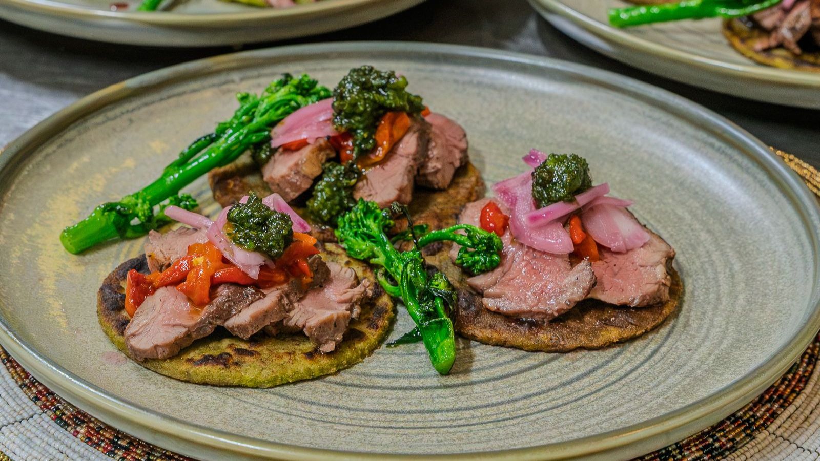Two corn tortillas topped with sliced steak, pickled onions, roasted peppers, pesto, and broccolini.