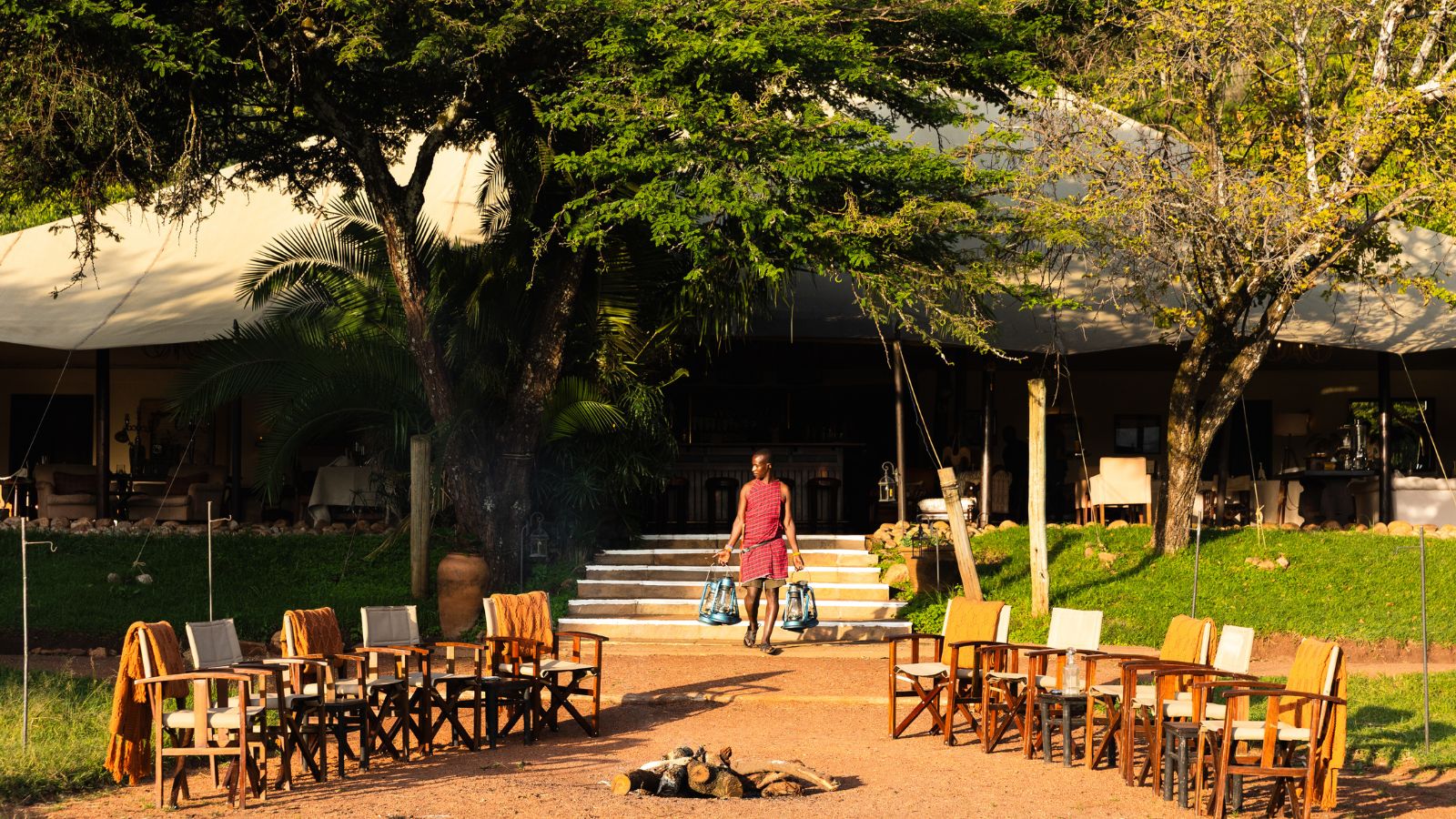 A person in traditional clothing walks toward a building carrying two lanterns, with chairs arranged in front of a fire pit.