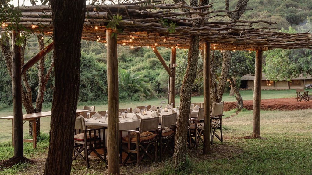 A long dining table set for a meal under a rustic wooden pergola with string lights at a Kenyan safari camp, located in a grassy outdoor area.