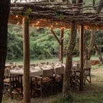 A long dining table set for a meal under a rustic wooden pergola with string lights at a Kenyan safari camp, located in a grassy outdoor area.