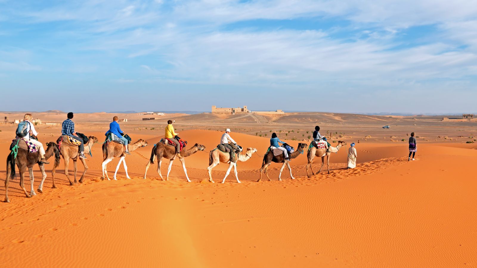 A group of people riding camels across orange sand dunes toward a building in the distance.