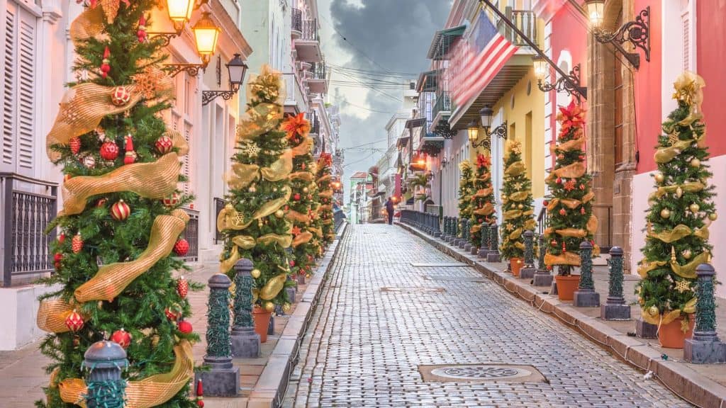 A cobblestone street in Old San Juan during a Puerto Rico Christmas, lined with decorated trees, colorful colonial buildings, and an American flag.