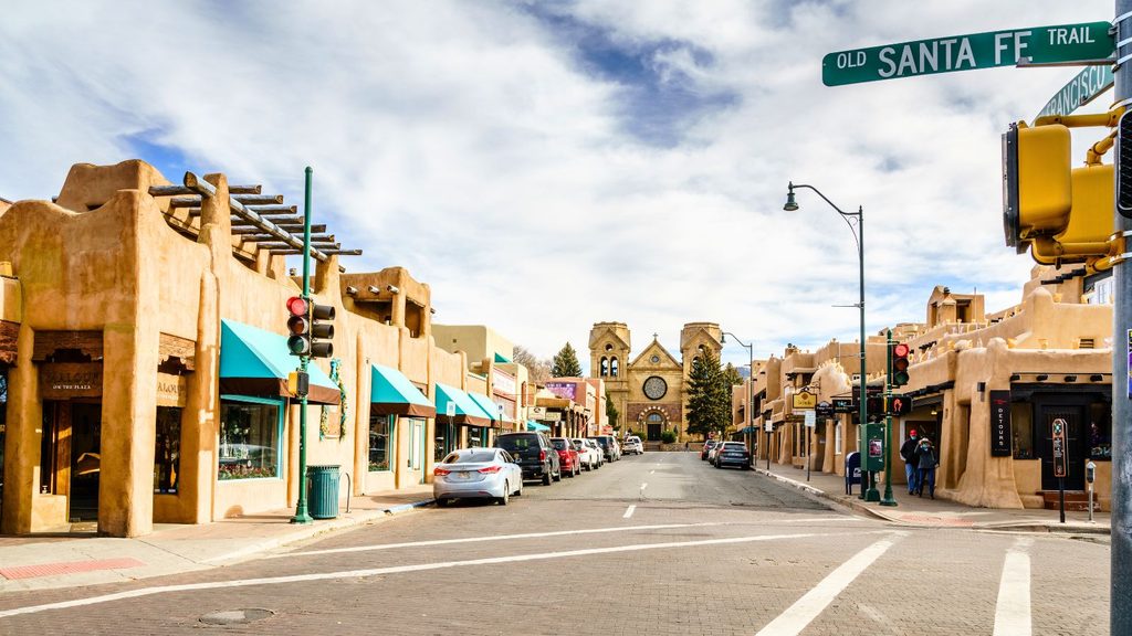 A street view of Old Santa Fe Trail, showing adobe buildings, a cathedral in the distance, and street signs under a blue sky.